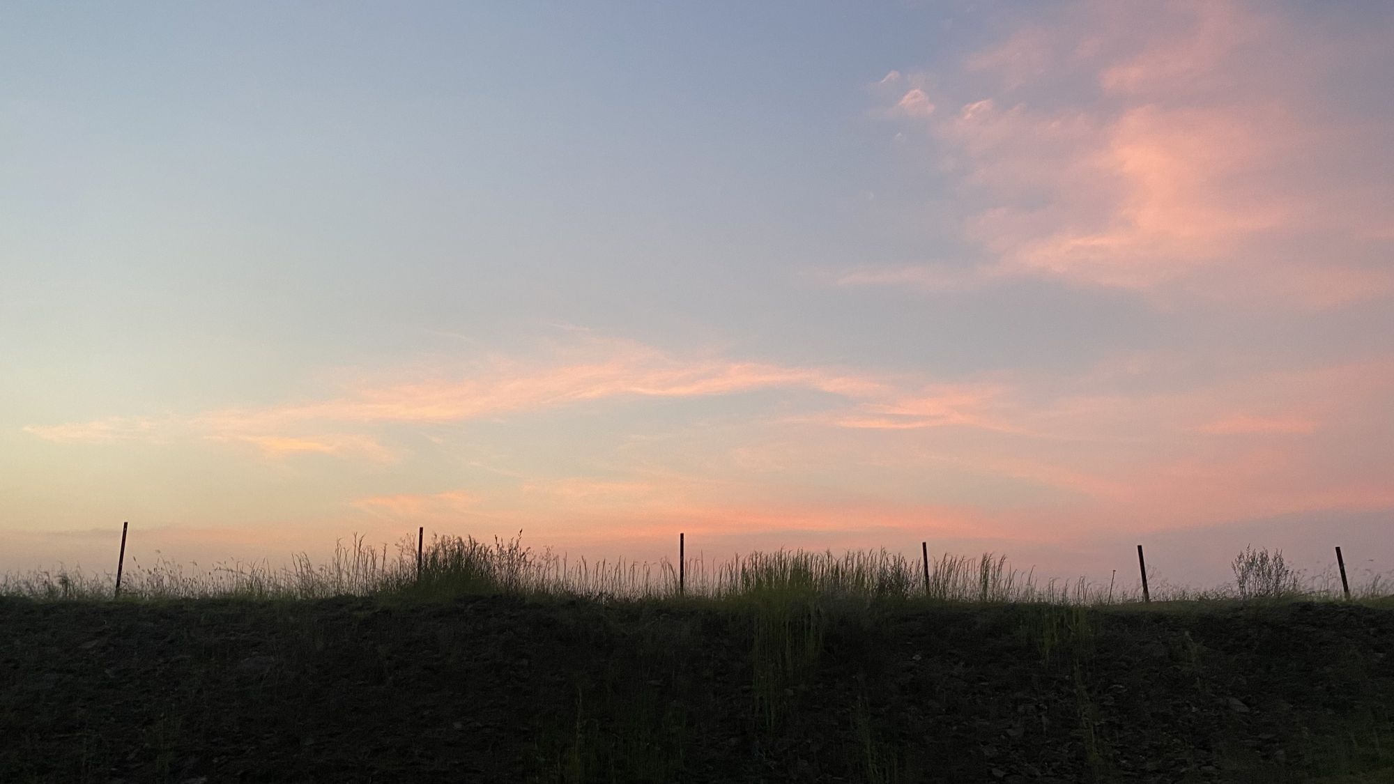Pink clouds in a blue evening sky above a dark ridge and fence line
